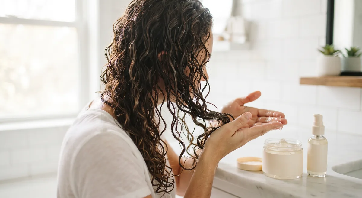 Mani che strizzano il gel per capelli ricci su ricci bagnati con lo scrunch, accanto a un vasetto aperto senza etichetta