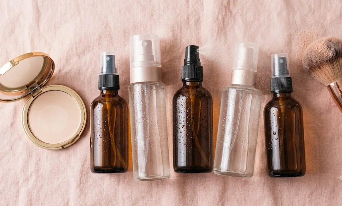 Cinque spray fissanti per il trucco di brand diversi allineati su piano di marmo chiaro con pennelli makeup
